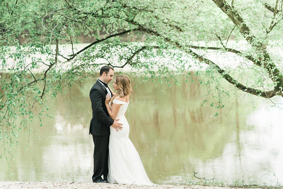Photo of bride and groom standing outside along the water's edge under a tree