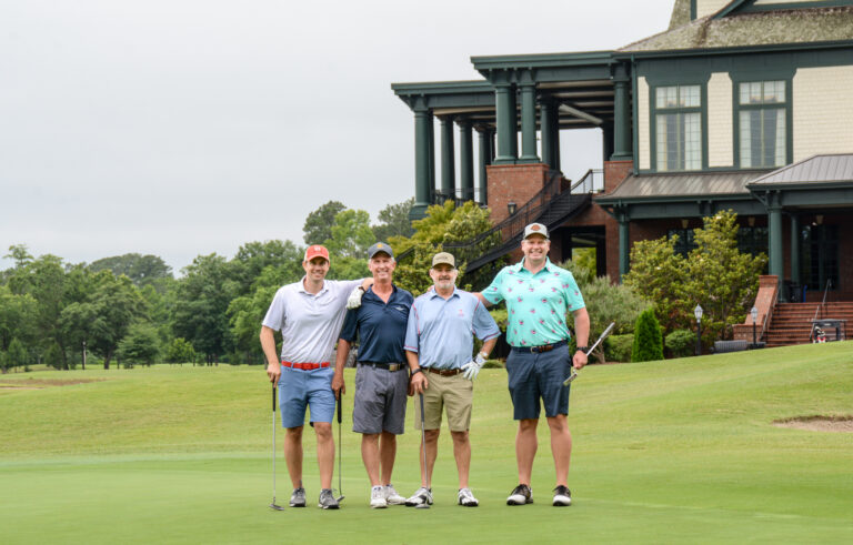 4 men golfers smiling with the background set as the clubhouse